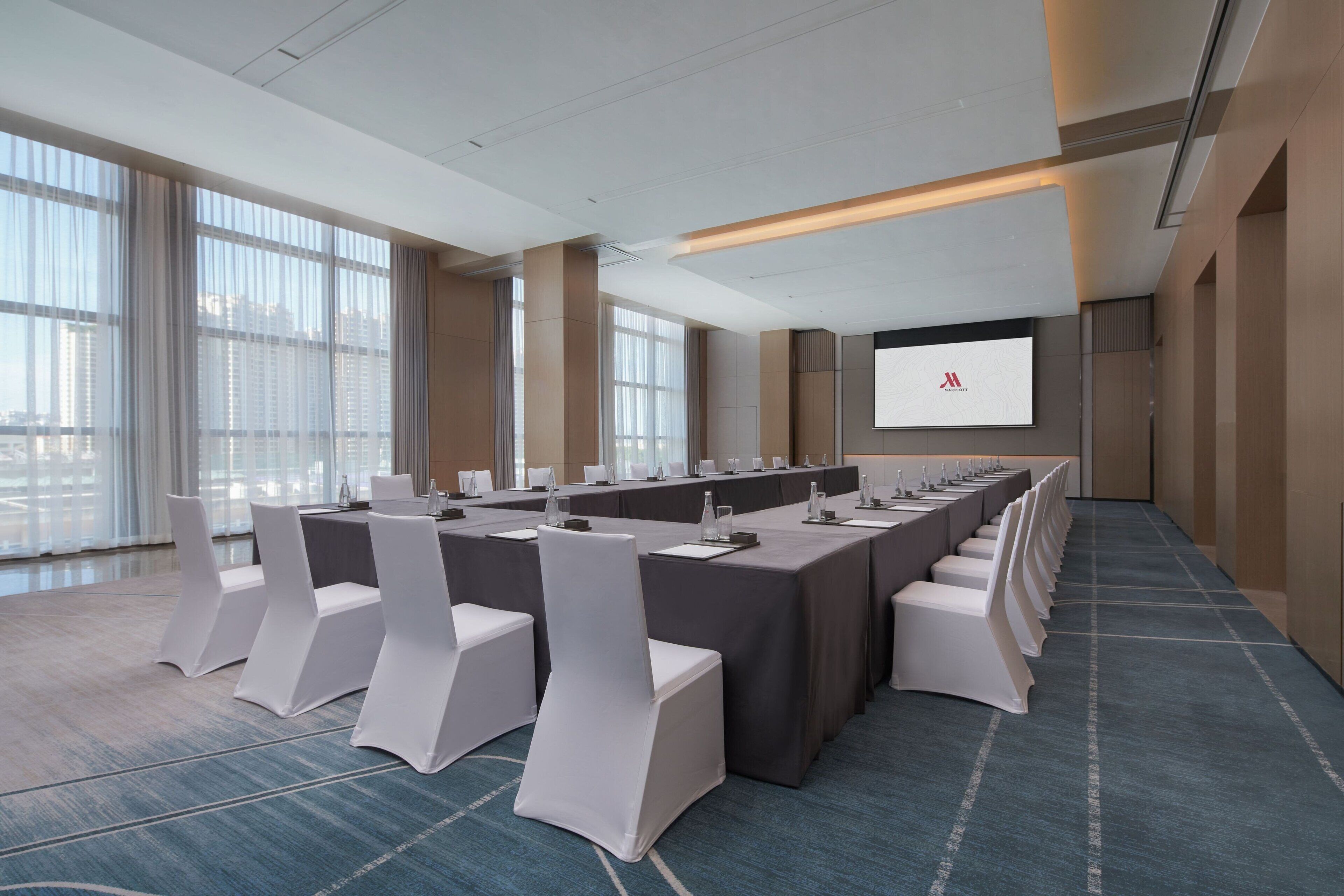 Un salón de conferencias con una larga mesa, sillas blancas y una pantalla para proyector.