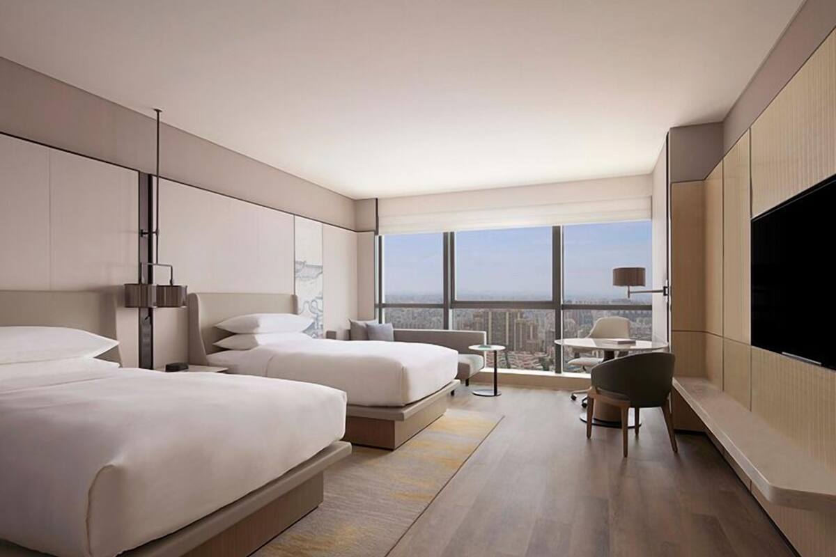A modern hotel room with two beds, a desk, a TV, and a large window offering a city view.