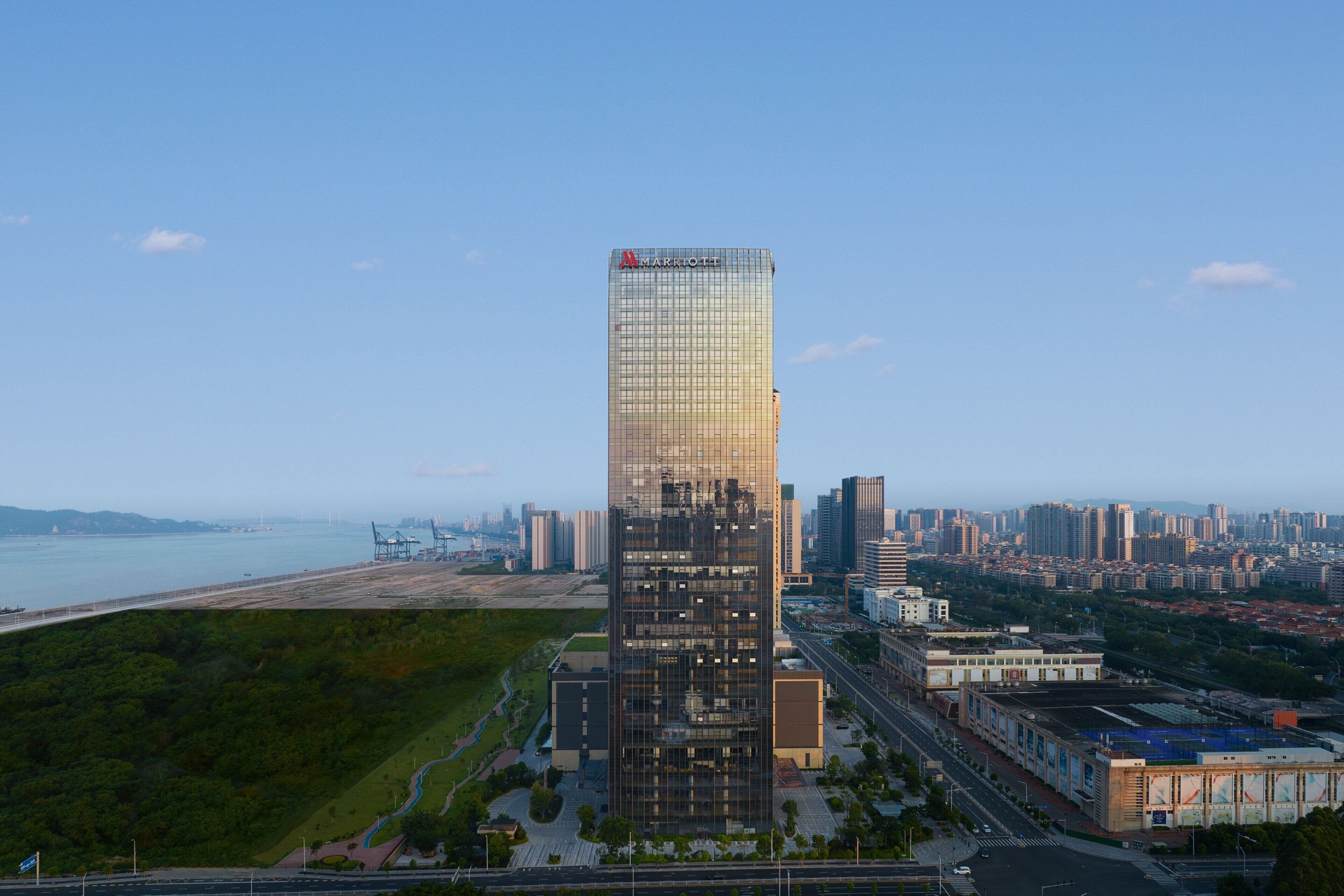 A modern high-rise hotel with a glass facade, located in an urban area with a view of the city skyline and a body of water in the background.