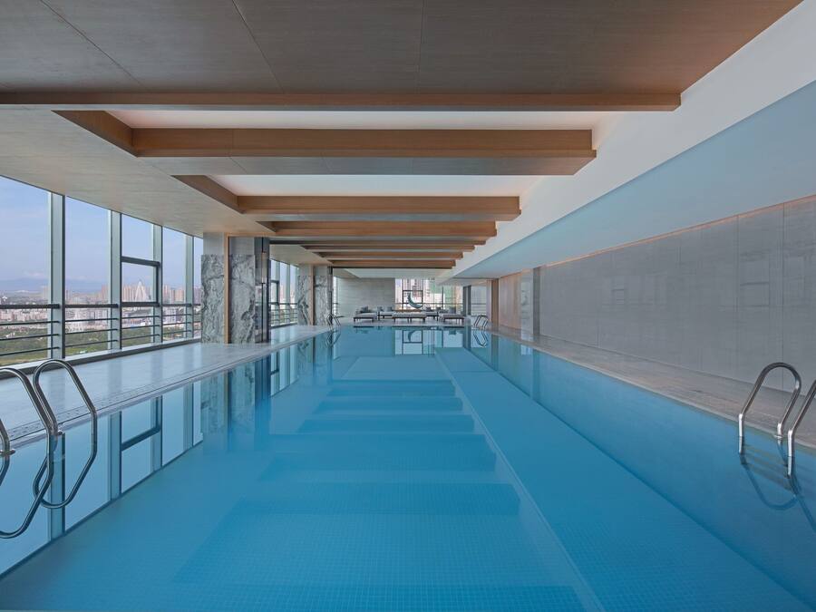 Une longue et étroite piscine intérieure, avec une eau d’un bleu limpide, des poutres en bois au plafond et de grandes fenêtres offrant une vue sur la ville.