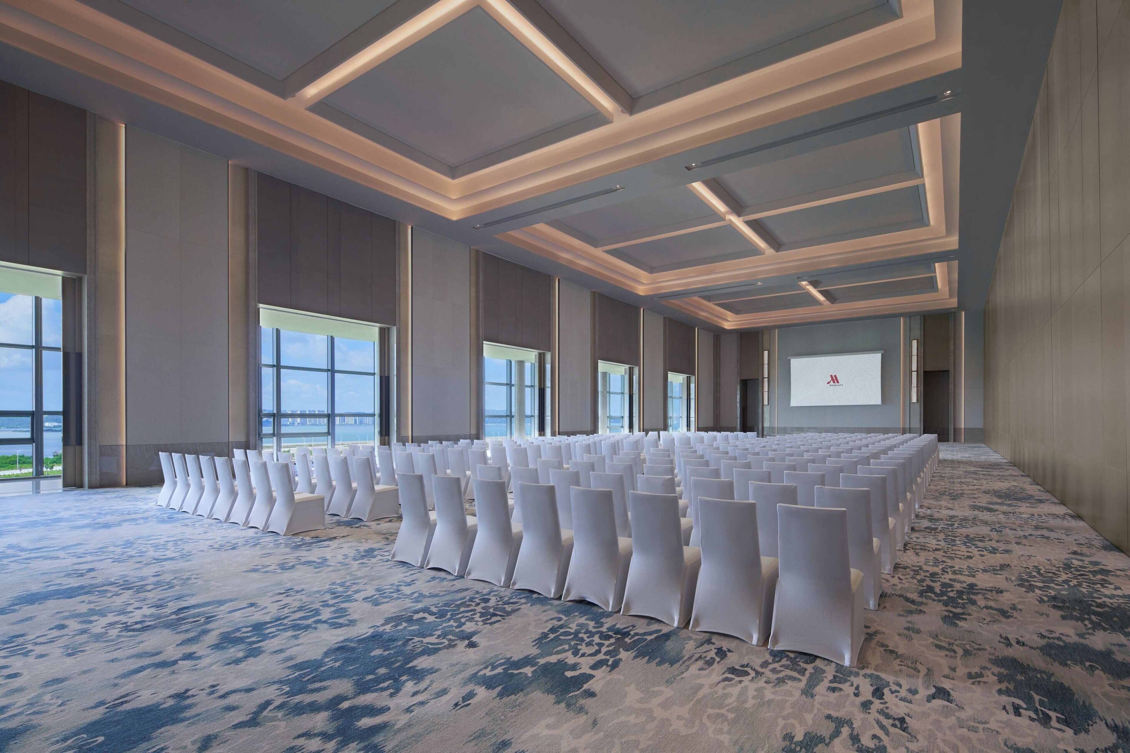Un salón de conferencias con filas de sillas blancas, ventanas amplias y suelo alfombrado.