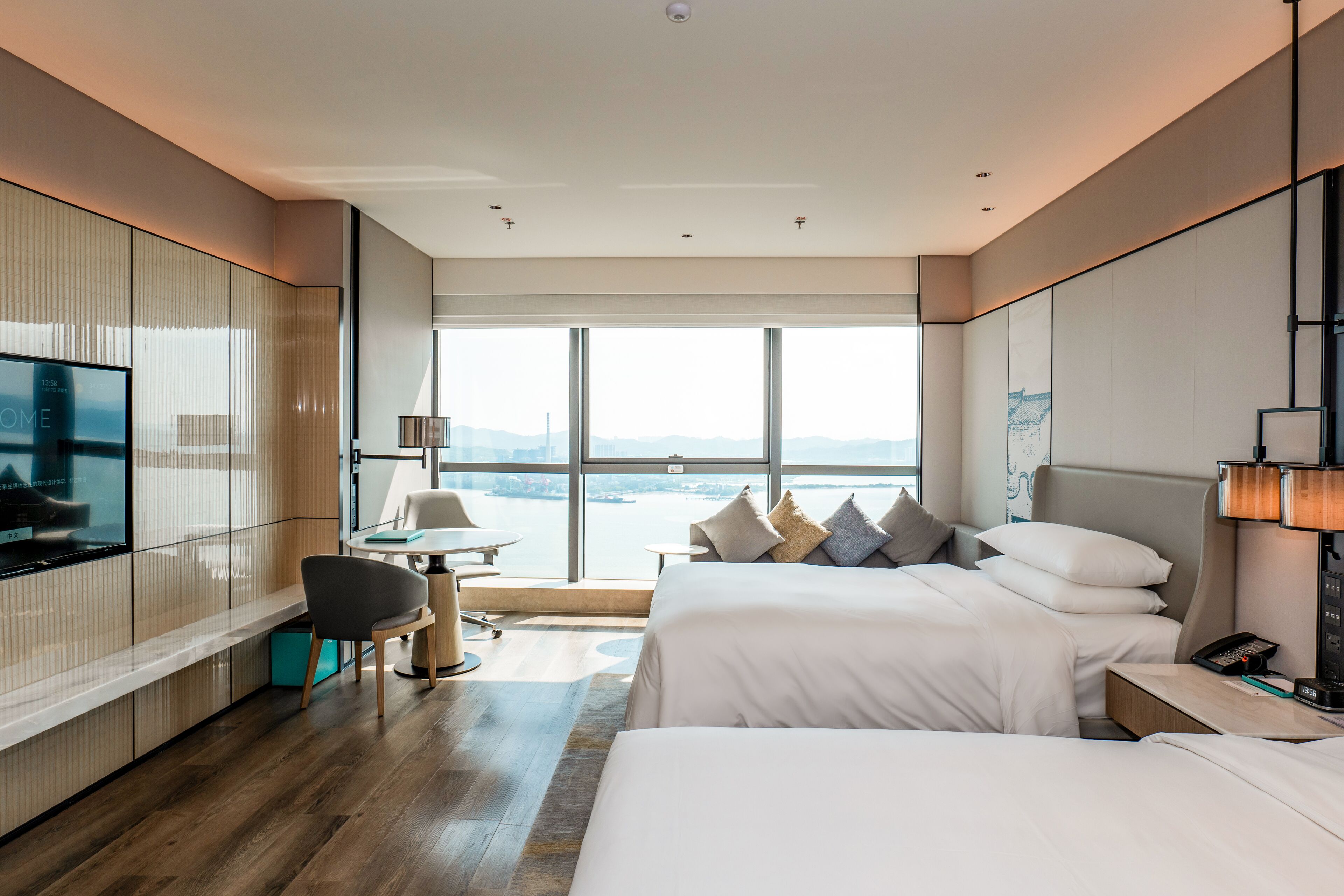 A modern hotel room with a large window, a TV, a desk, and two beds.