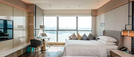 A modern hotel room with a large window, a TV, a desk, and two beds.
