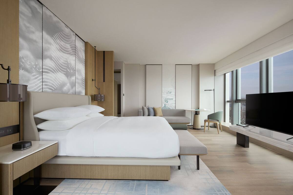 A modern hotel room with a large bed, a flat-screen TV, a desk, and a view of the city through large windows.