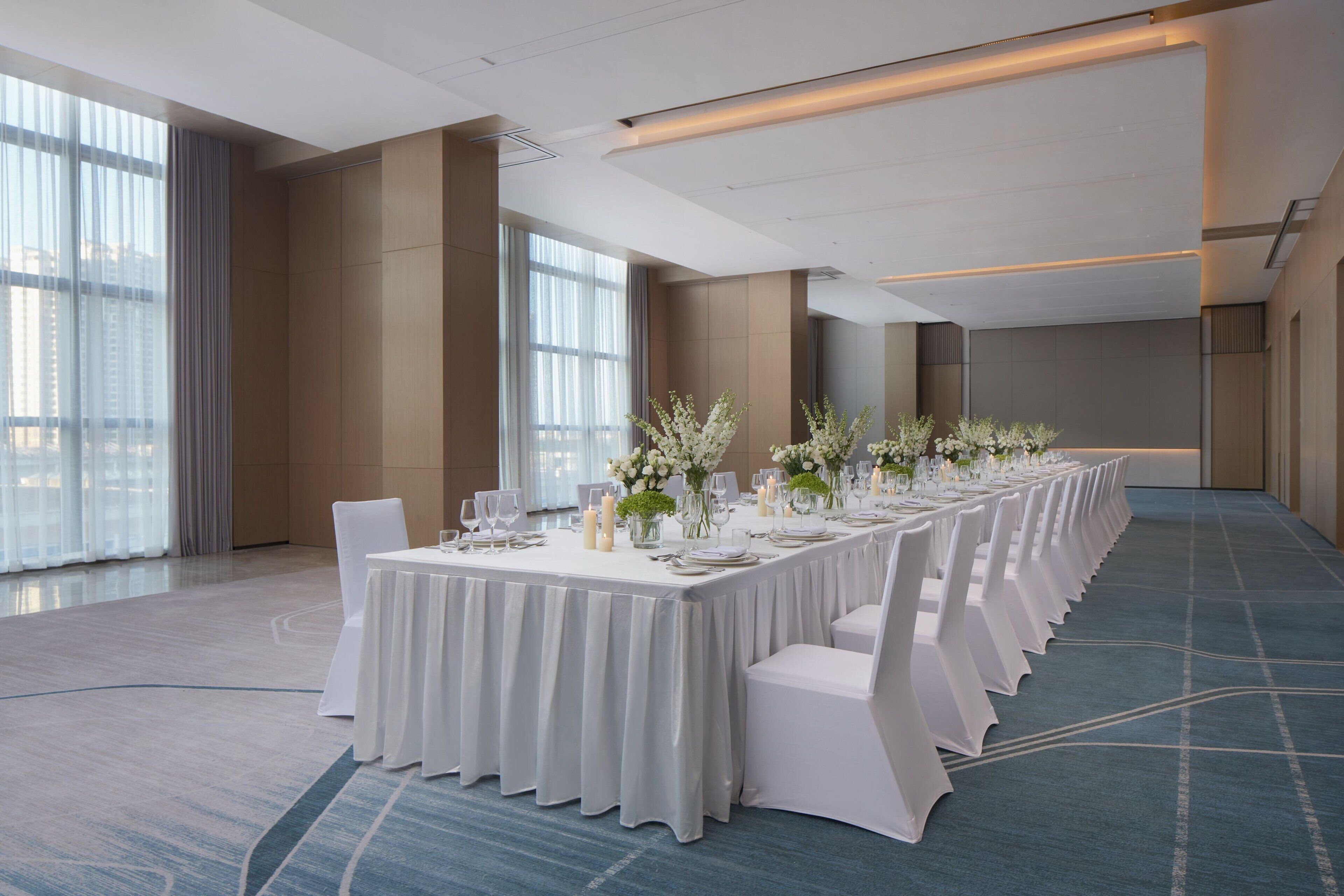 A long banquet table set with white tablecloths, chairs, and floral centerpieces in a spacious room with large windows and a carpeted floor.