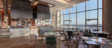 A modern restaurant with a large kitchen, dining area with tables and chairs, and large windows offering a city view.