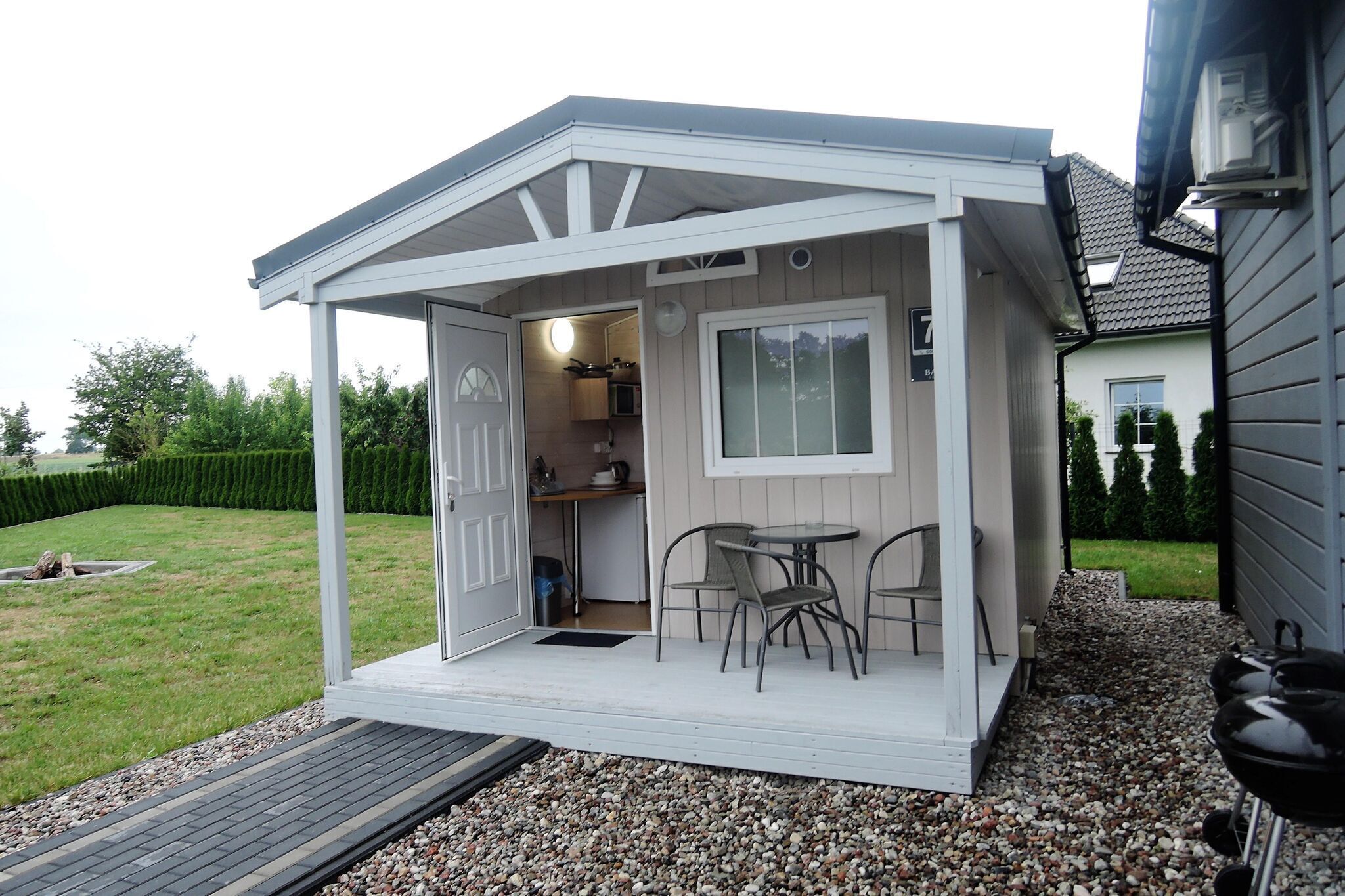 A small, white, covered outdoor kitchen area with a table and chairs, a door leading inside, and a grill outside.