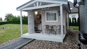 A small, white, covered outdoor kitchen area with a table and chairs, a door leading inside, and a grill outside.