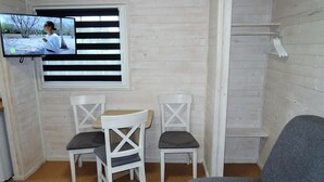 A room with a TV mounted on the wall, two white chairs with cushioned seats, a grey sofa, and a wooden shelf with a towel rack.