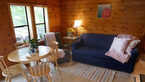 A cozy cabin interior with a wooden dining table, chairs, a blue sofa, and a small side table with a lamp and flowers.