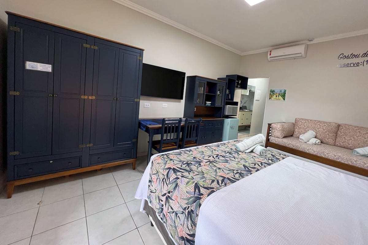 A hotel room with a large wardrobe, a desk with a chair, a bed with a floral bedspread, and a sofa with towels.