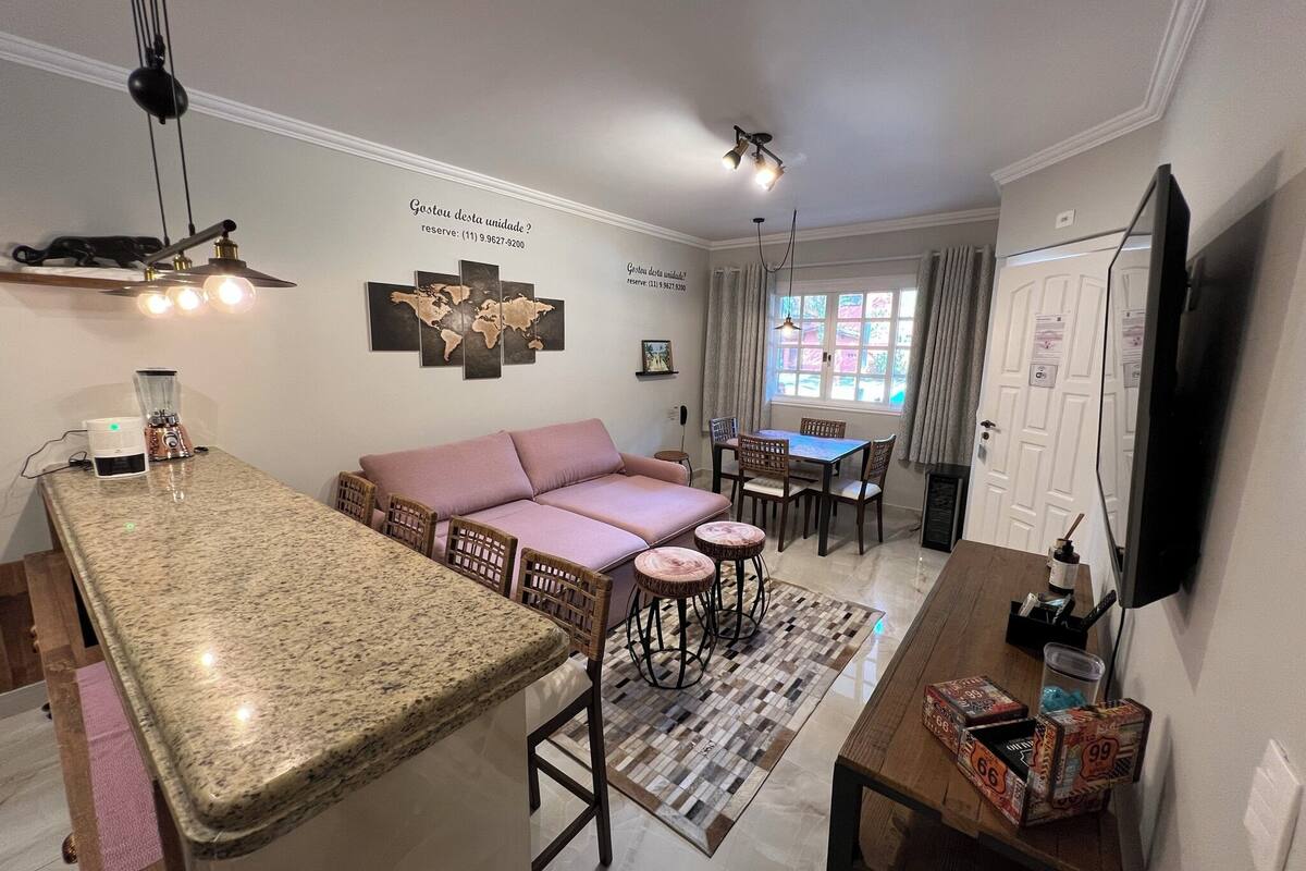 A modern living room with a kitchen area, a dining table, and a sofa. There is a world map on the wall and a window with curtains.