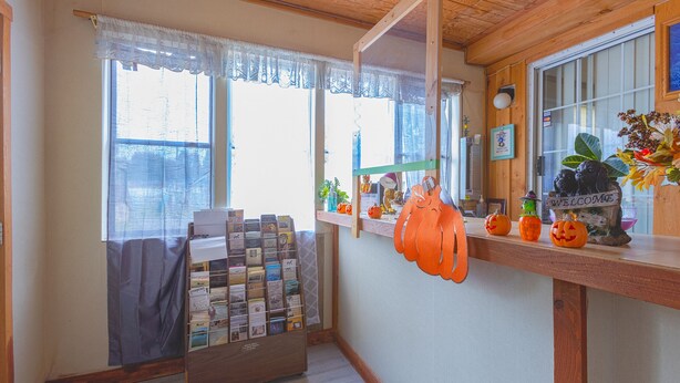 Una habitación con techo de madera, una ventana con cortina de encaje, un tablón de anuncios con tarjetas y una estantería con calabazas y un cartel de bienvenida.
