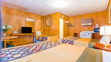A hotel room with wooden walls, a TV, a desk with a computer, two beds with colorful bedspreads, a mirror, a kitchen area with a microwave, and a coffee maker.