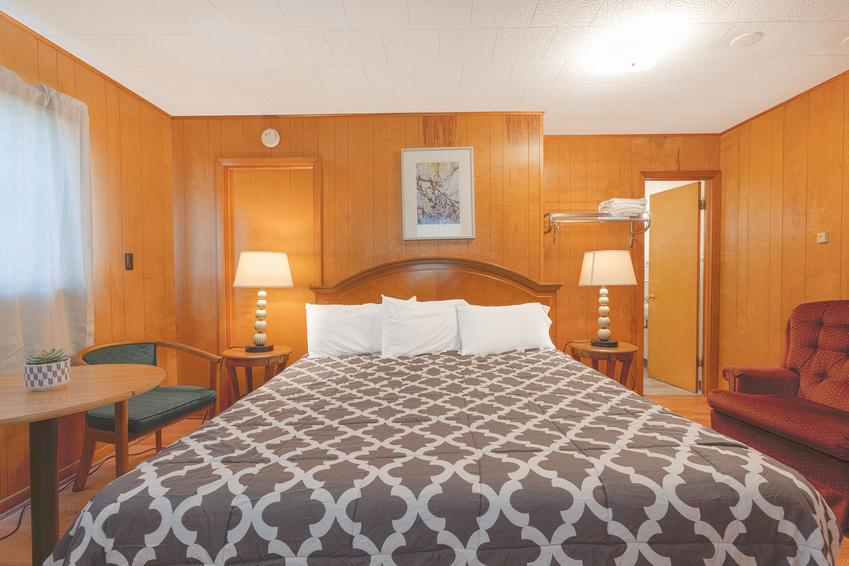 A hotel room with a large bed, two bedside lamps, a wooden headboard, a small table with a chair, and a red armchair.