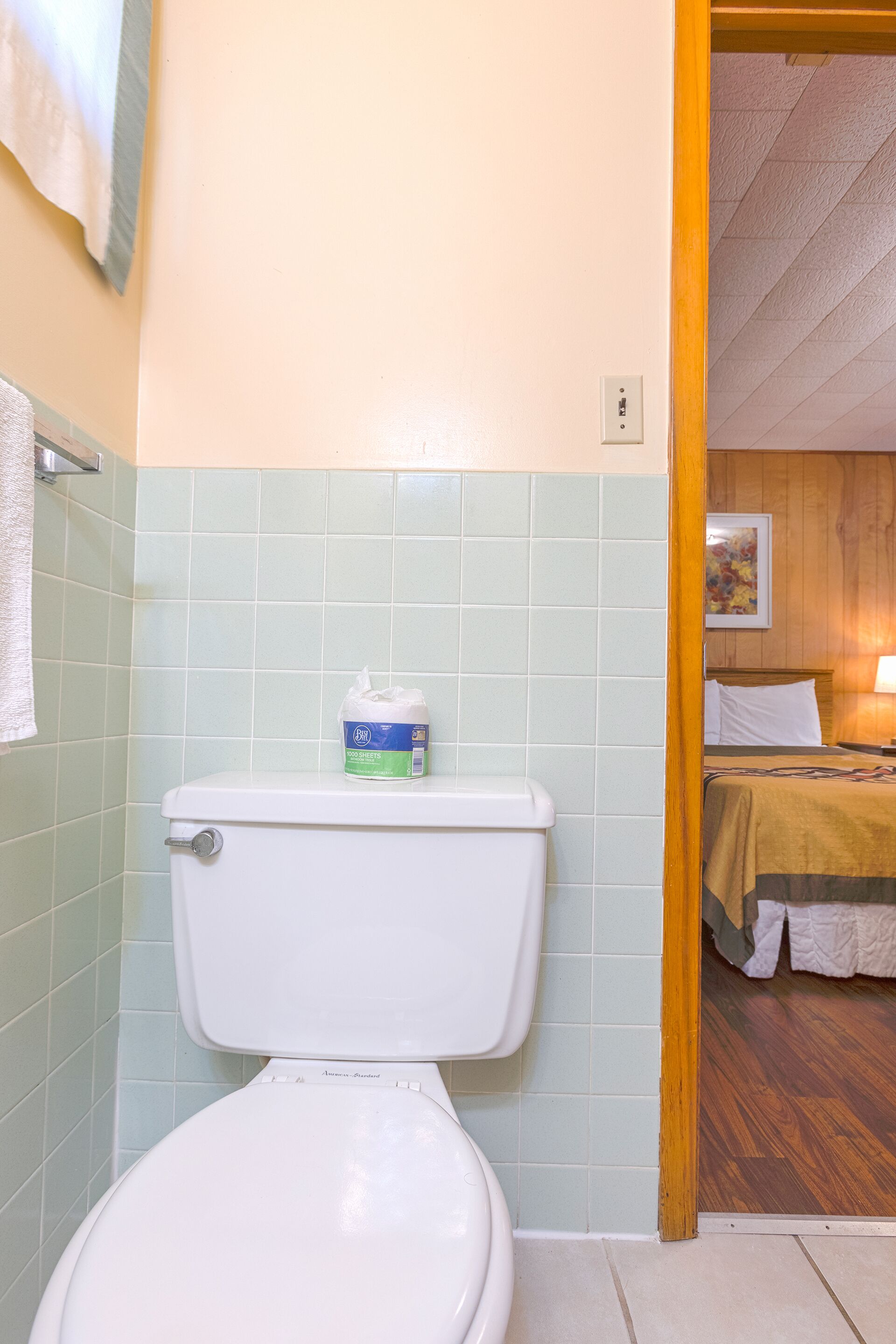 A bathroom with a toilet, green tiles, a white towel, and a wooden floor leading to a bedroom with a bed and a painting.