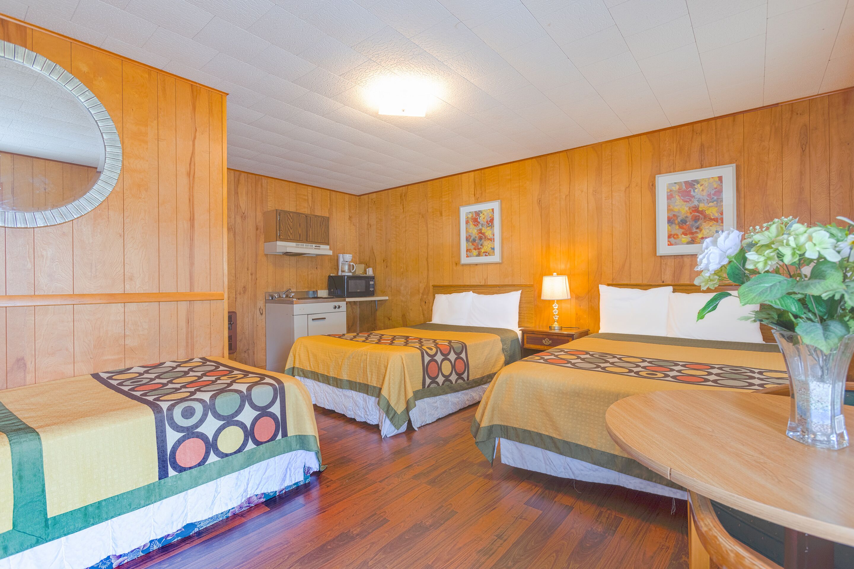 A hotel room with two beds, a wooden floor, a mirror, a desk, a lamp, and a vase with flowers.