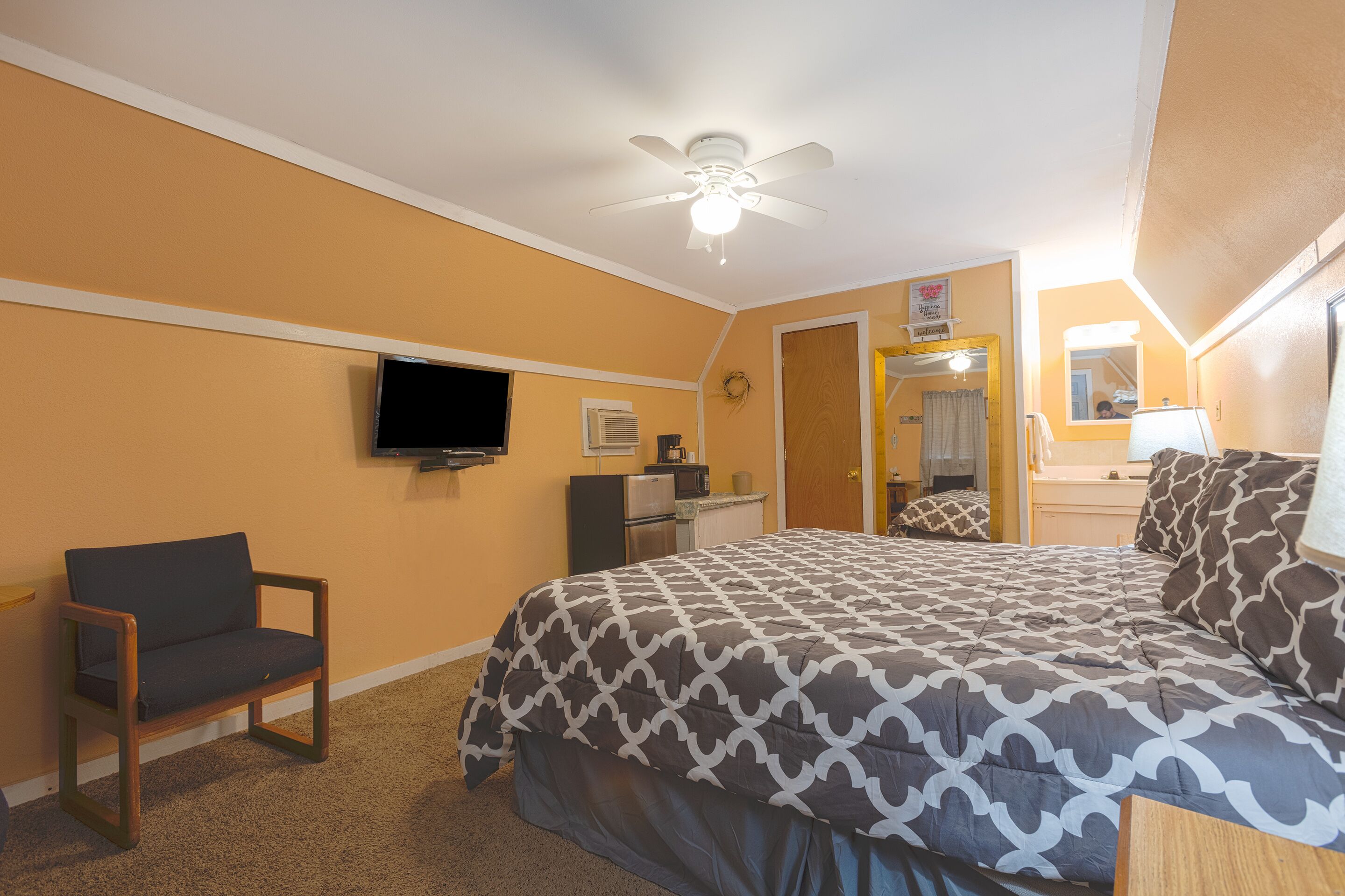 A hotel room with a bed, a chair, a television, a ceiling fan, and a bathroom with a mirror and sink.