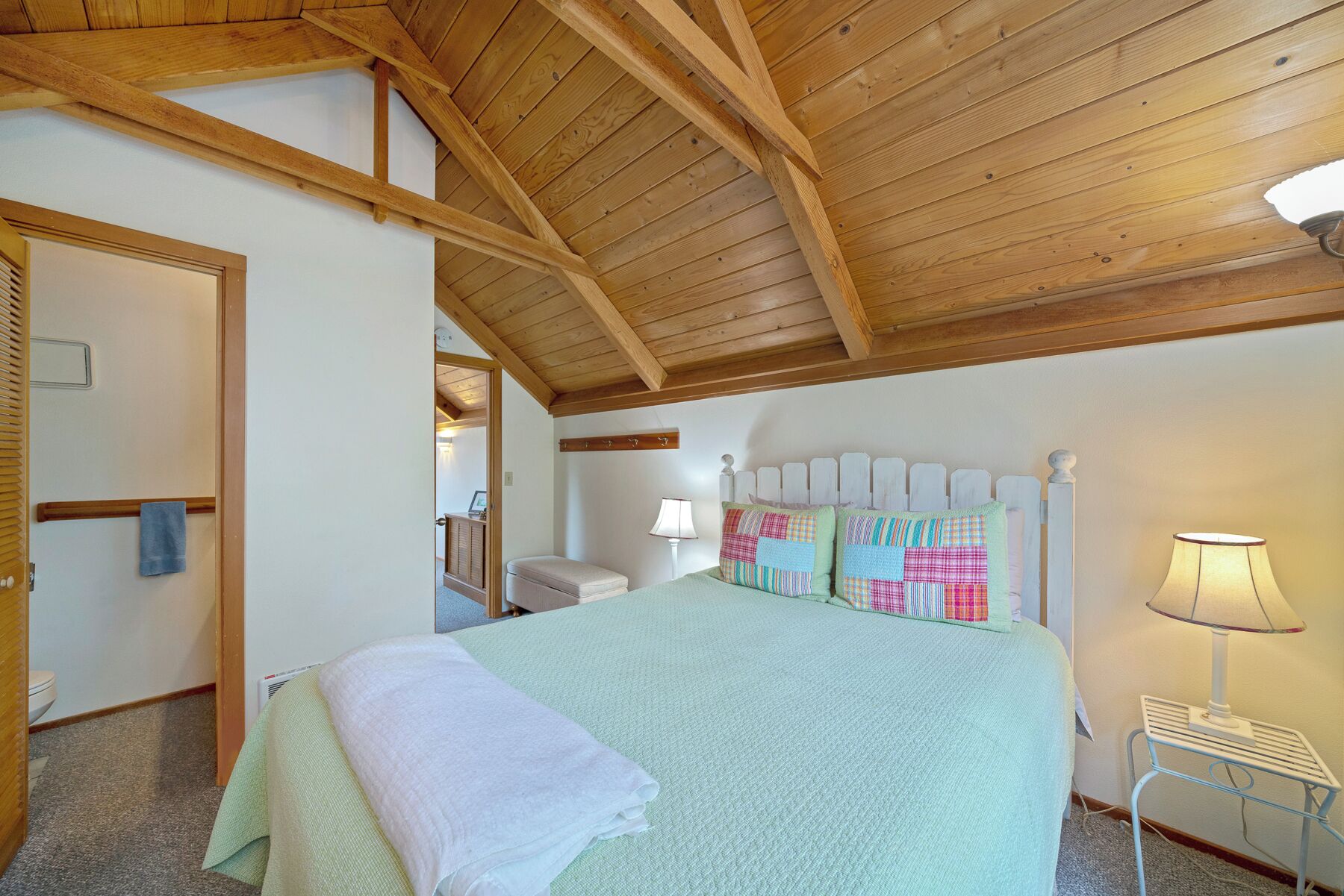 Un dormitorio con un techo alto de madera, una cama con colcha, una lámpara de noche y una mesita.