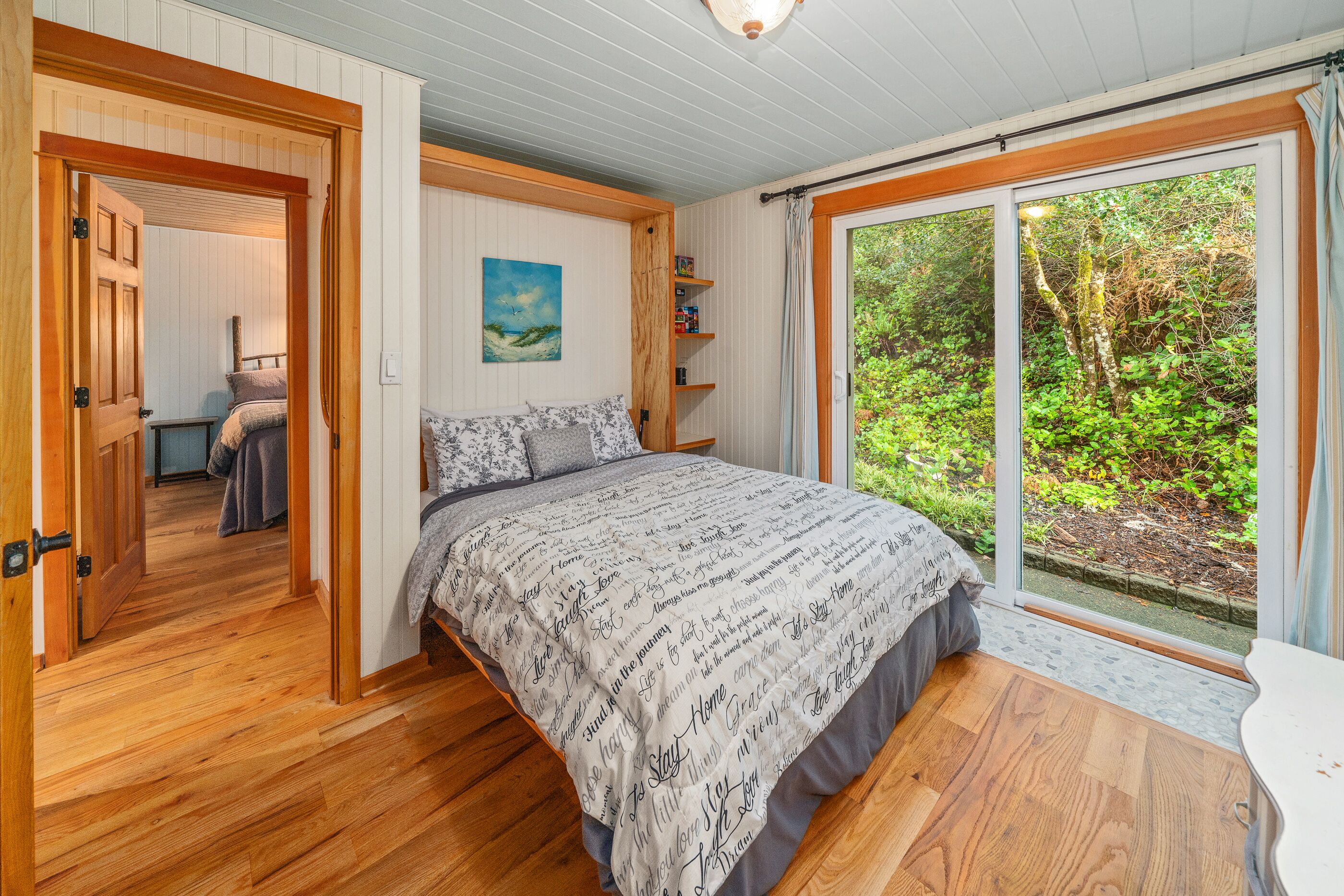 Een slaapkamer met een groot bed, houten vloeren, een raam met uitzicht op groen en een deur naar een andere kamer.