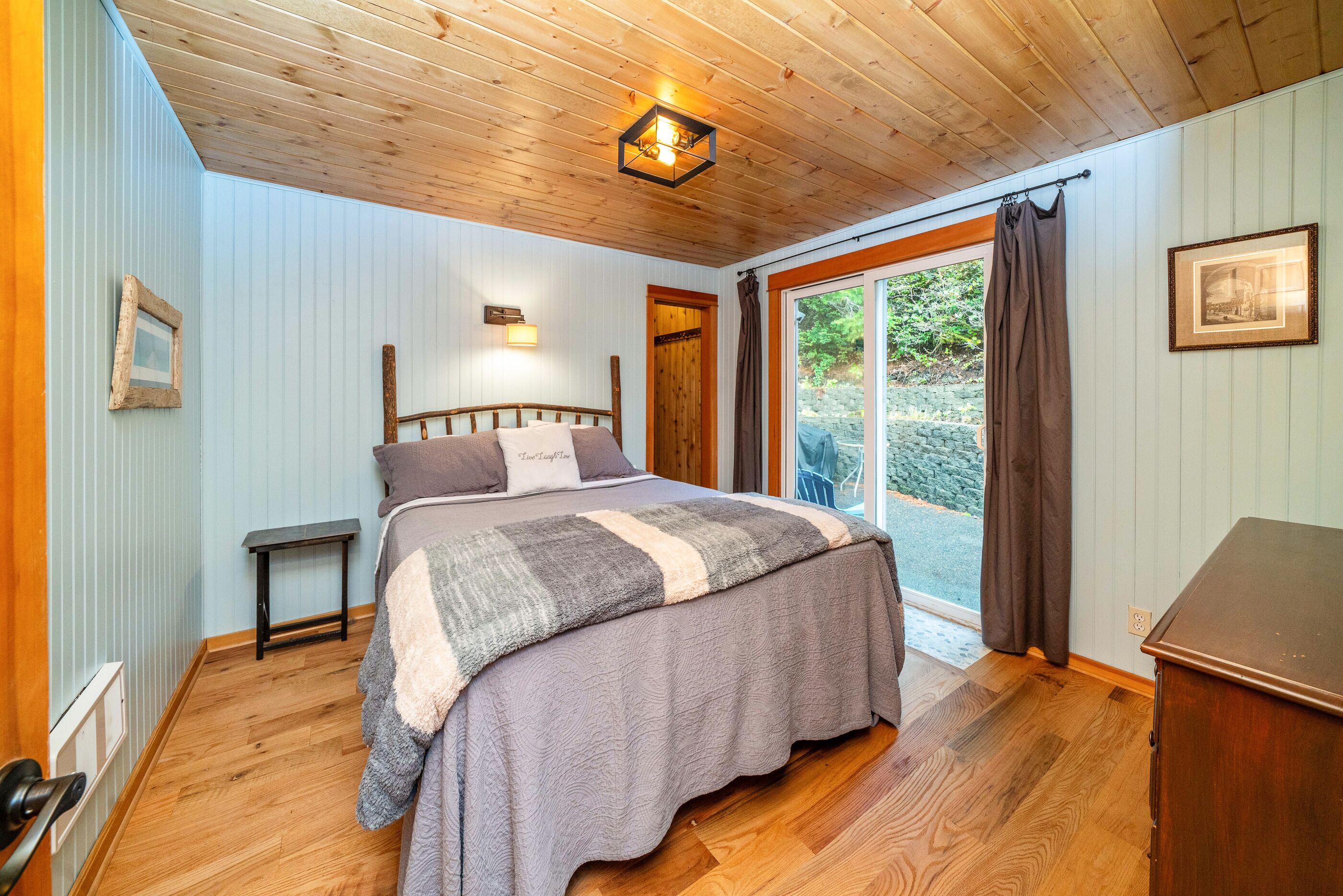 A bedroom with a wooden ceiling, a bed with a grey and white striped blanket, a bedside table, and a view of the outdoors through a glass door.