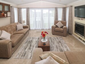 A spacious living room with a beige sofa, a brown armchair, and a coffee table with a vase of flowers. A flat-screen TV is mounted on the wall, and there is a fireplace on the right side. - Gwel Eryri (Gaerwen)