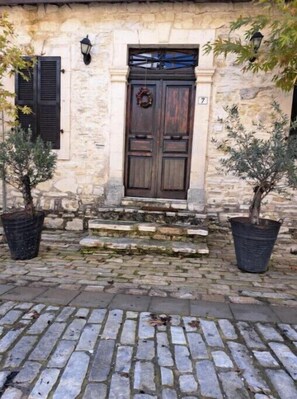 Un edificio de piedra con puerta y contraventanas de madera, plantas en macetas a ambos lados, y un camino adoquinado que lleva a la entrada.
