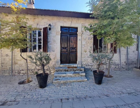 A stone building with a dark wooden door, two windows with shutters, and a cobblestone courtyard.