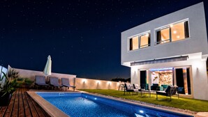 Una casa moderna con piscina, sillas de descanso y un espacio exterior bien iluminado.