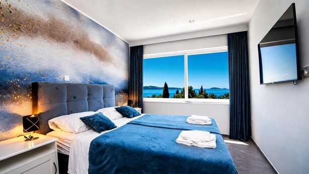 Una habitación de hotel moderna con una cama grande, una mesita de noche, una lámpara, un televisor y una ventana con vista al mar.