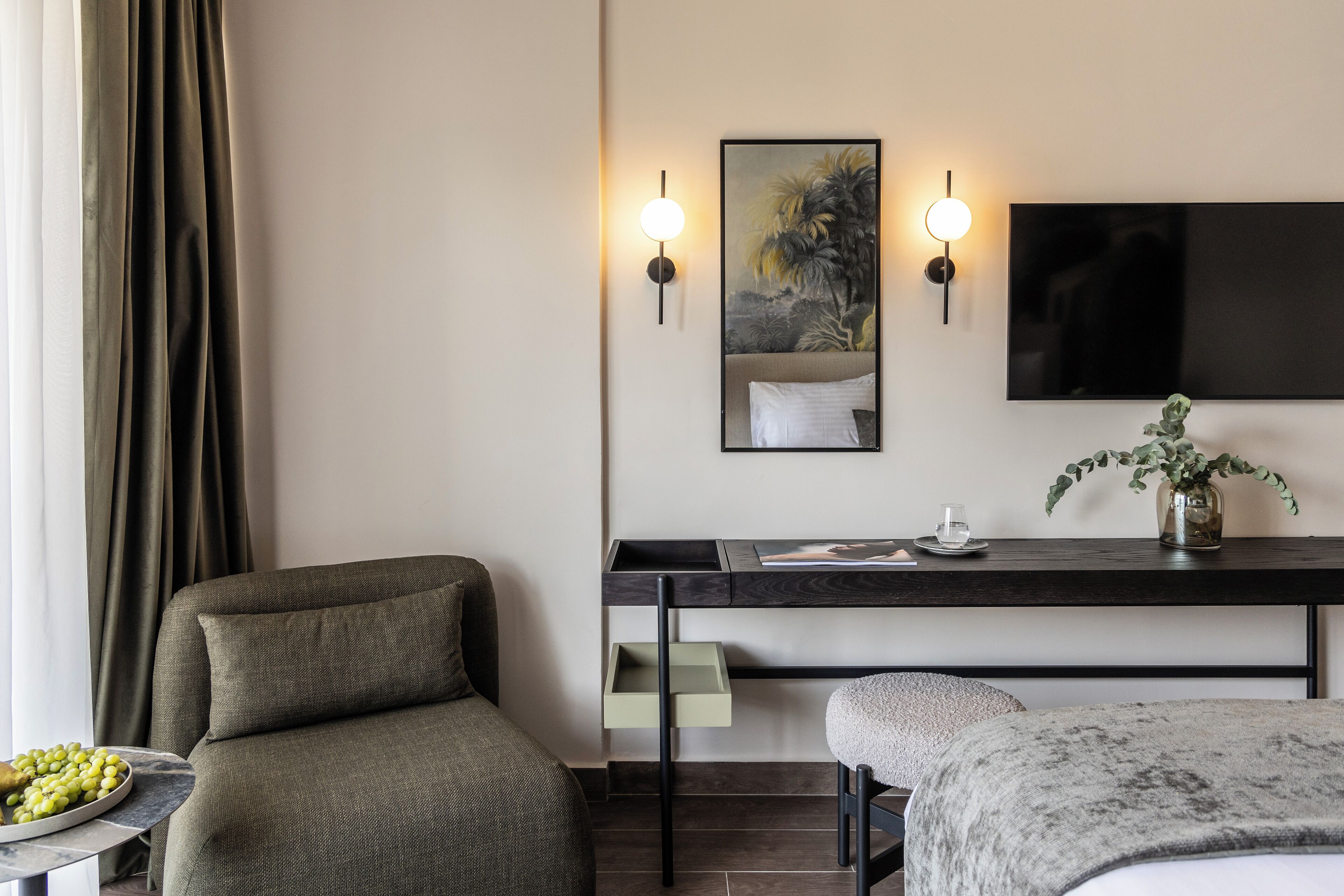 A modern hotel room with a sofa, a small table with a bowl of fruit, a desk with a flat-screen TV, and a wall-mounted light fixture.