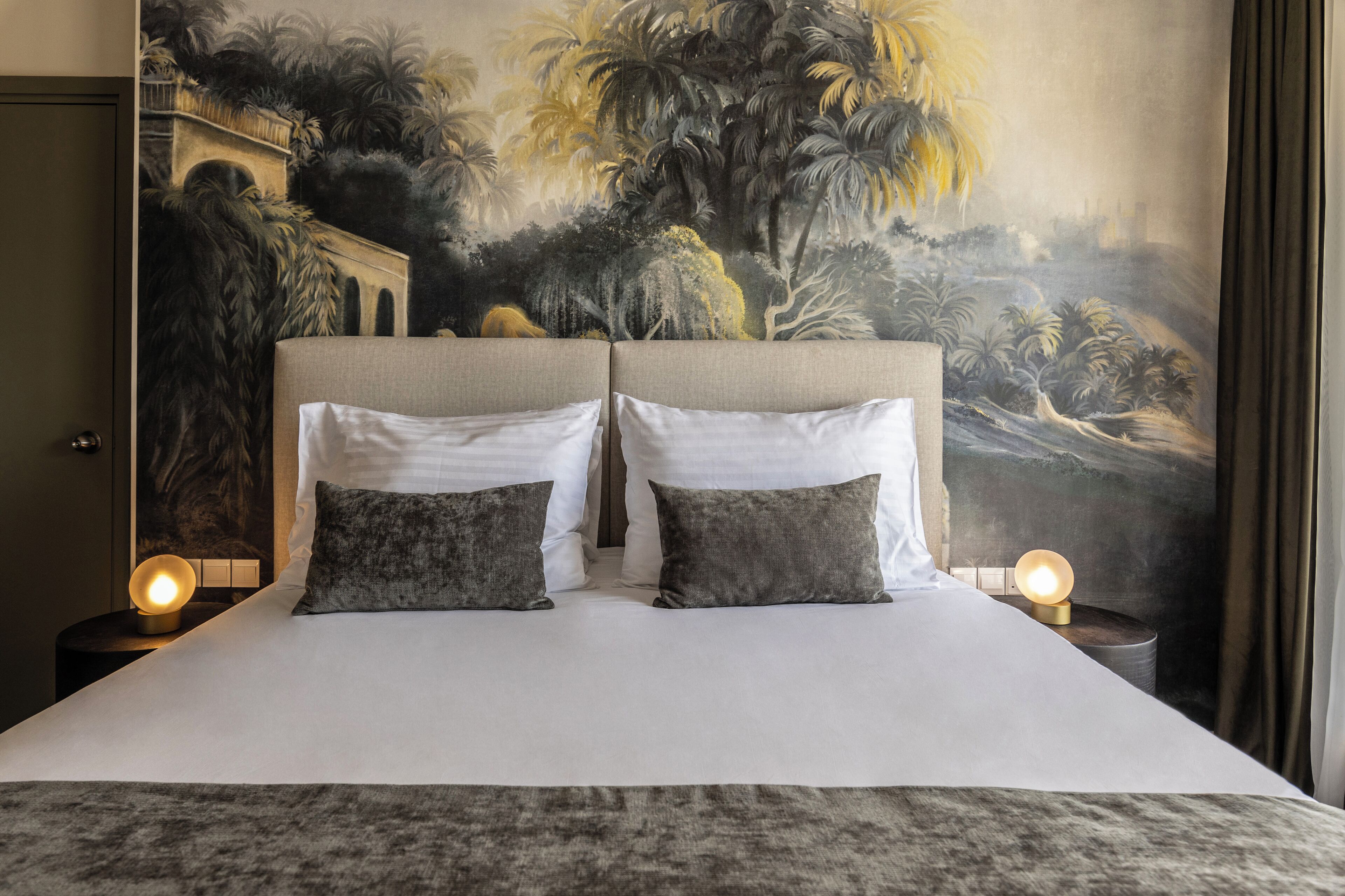 A bed with white linens and two pillows, a nightstand with a lamp, and a large mural on the wall depicting a tropical landscape.