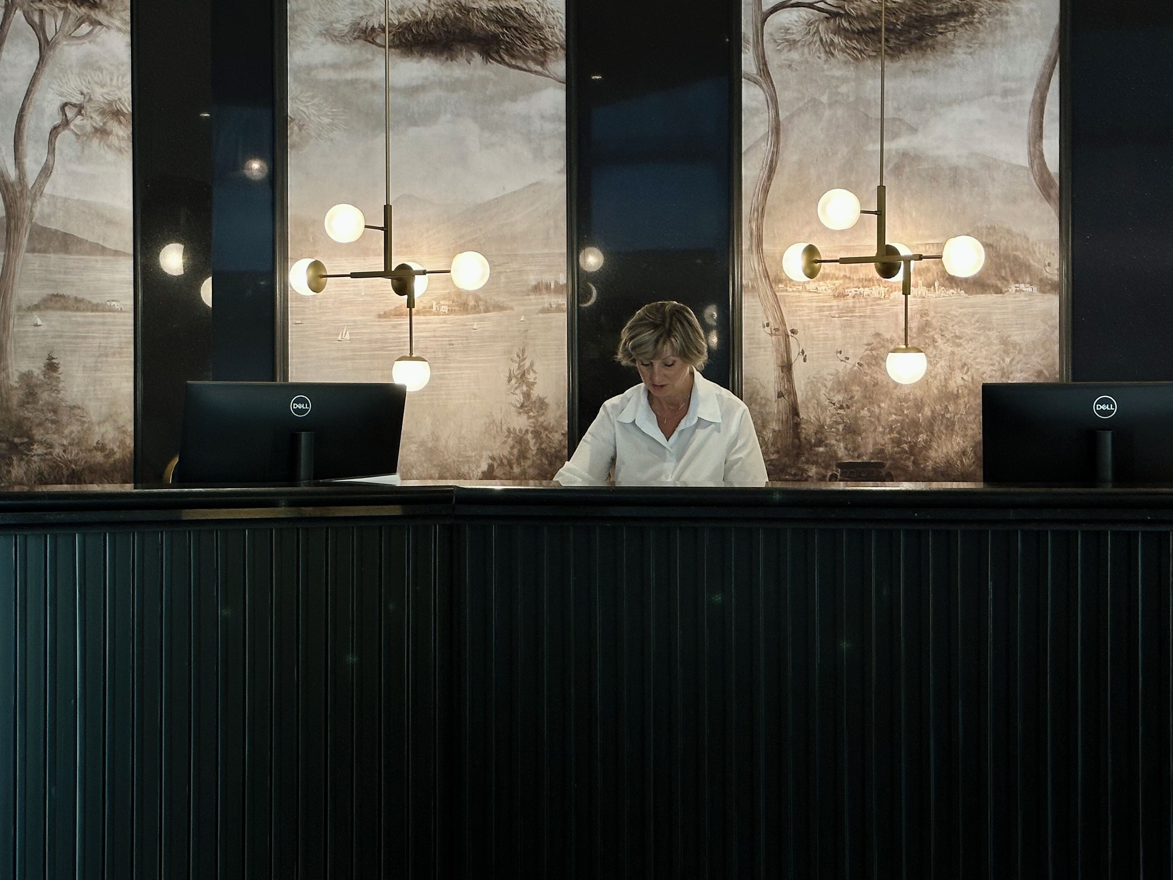 A reception desk with two Dell computers, a large landscape painting, and a hanging light fixture.