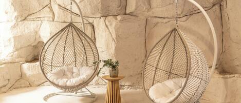 Dos sillones colgantes tipo huevo con cojines, una mesita de madera con un florero, y una pared con textura.