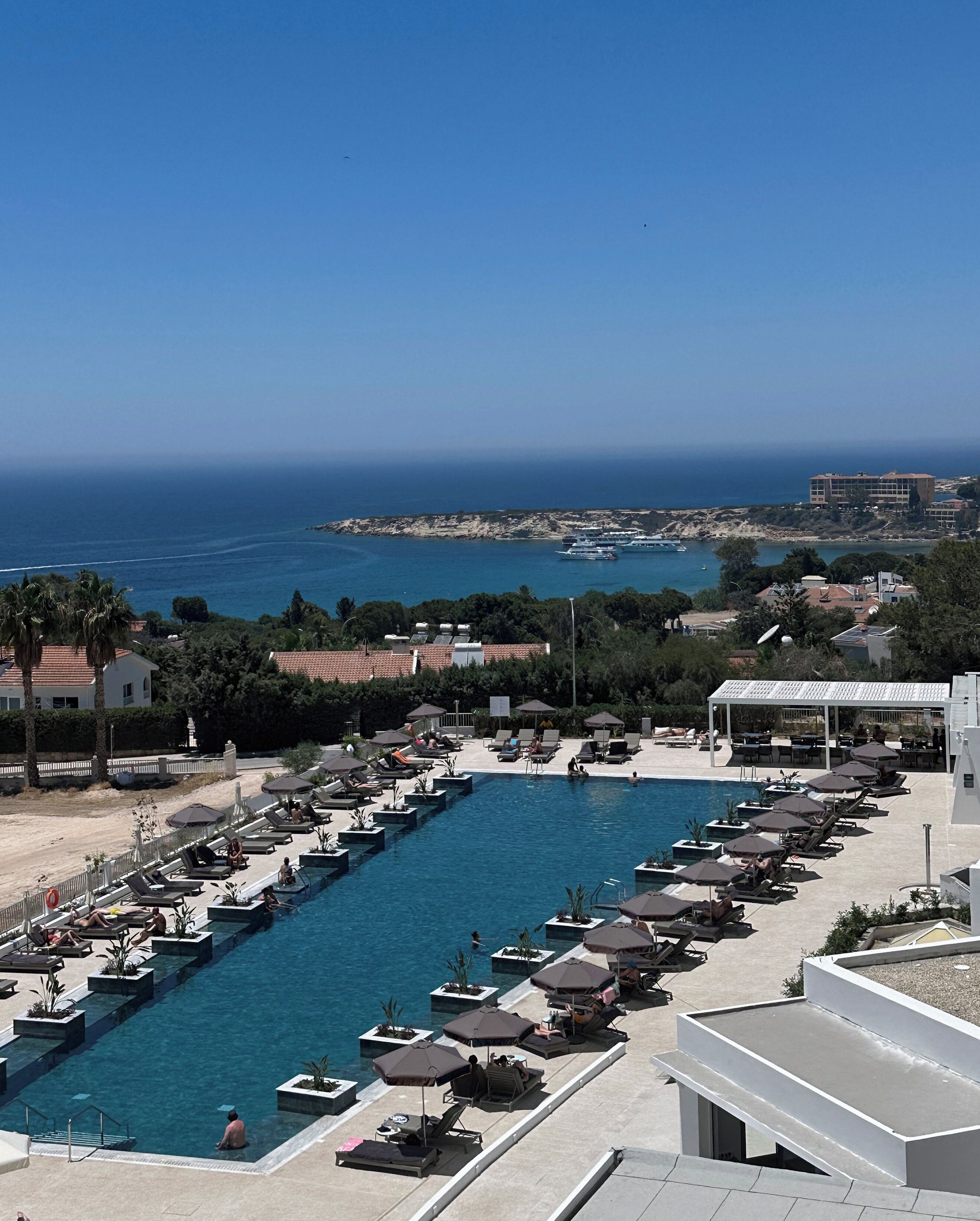 Un espace piscine doté de nombreuses chaises longues et de parasols, entouré d’un jardin aménagé et offrant une vue sur la côte.