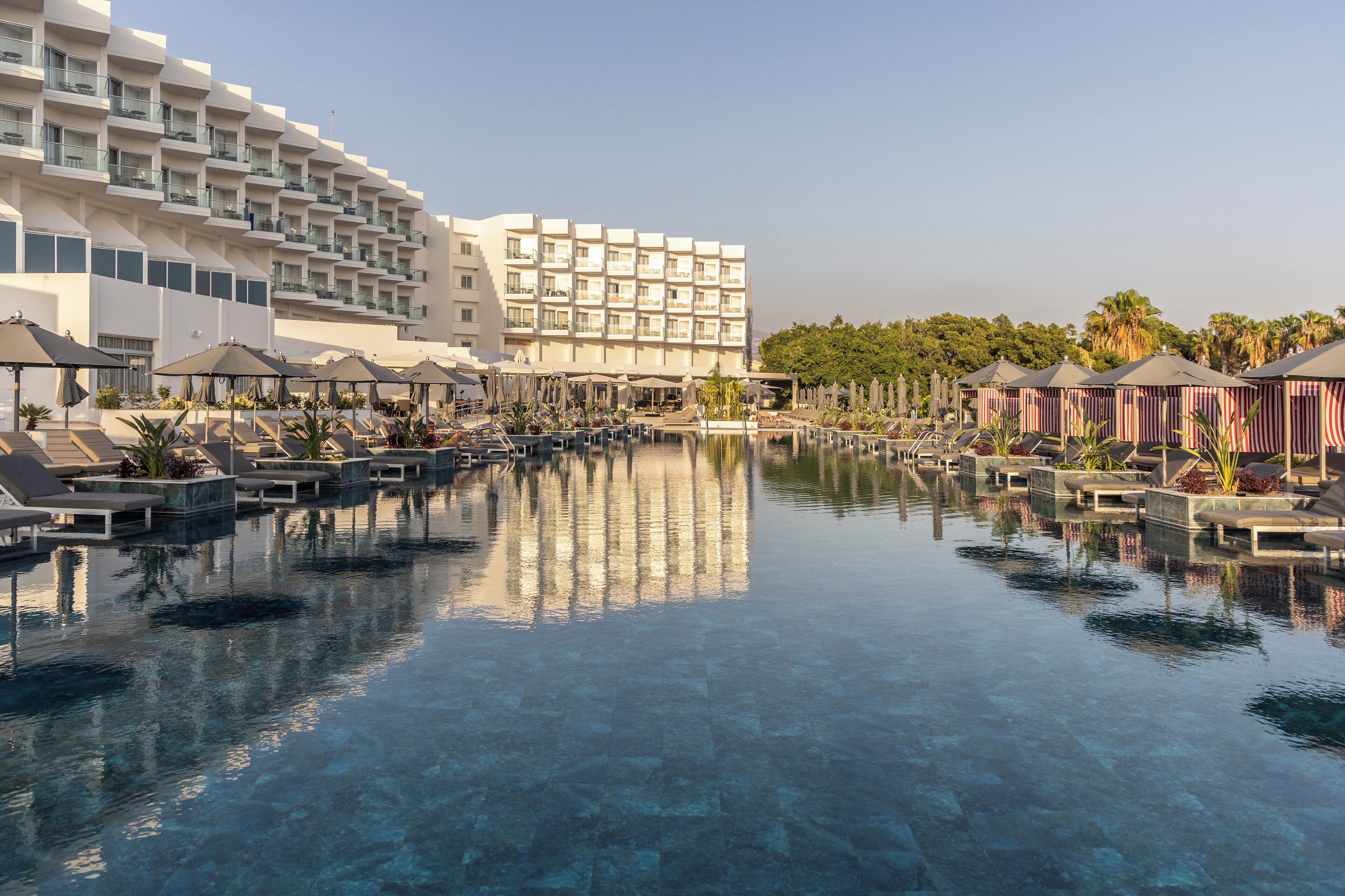 Un hotel con una piscina amplia, sillas de descanso, sombrillas y un edificio moderno con balcones.
