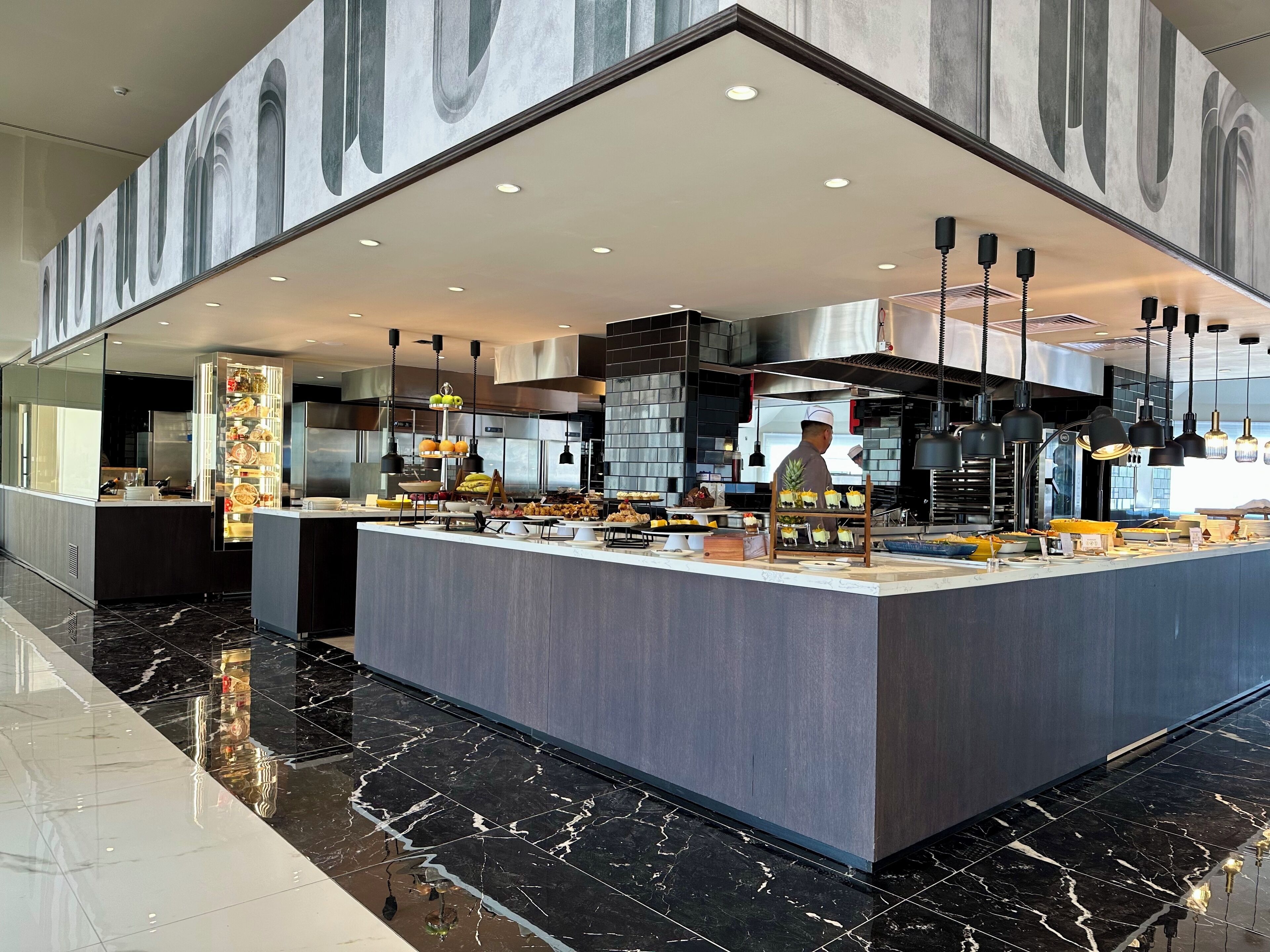 A modern kitchen with a marble floor, a central island with food displays, and a chef preparing food.