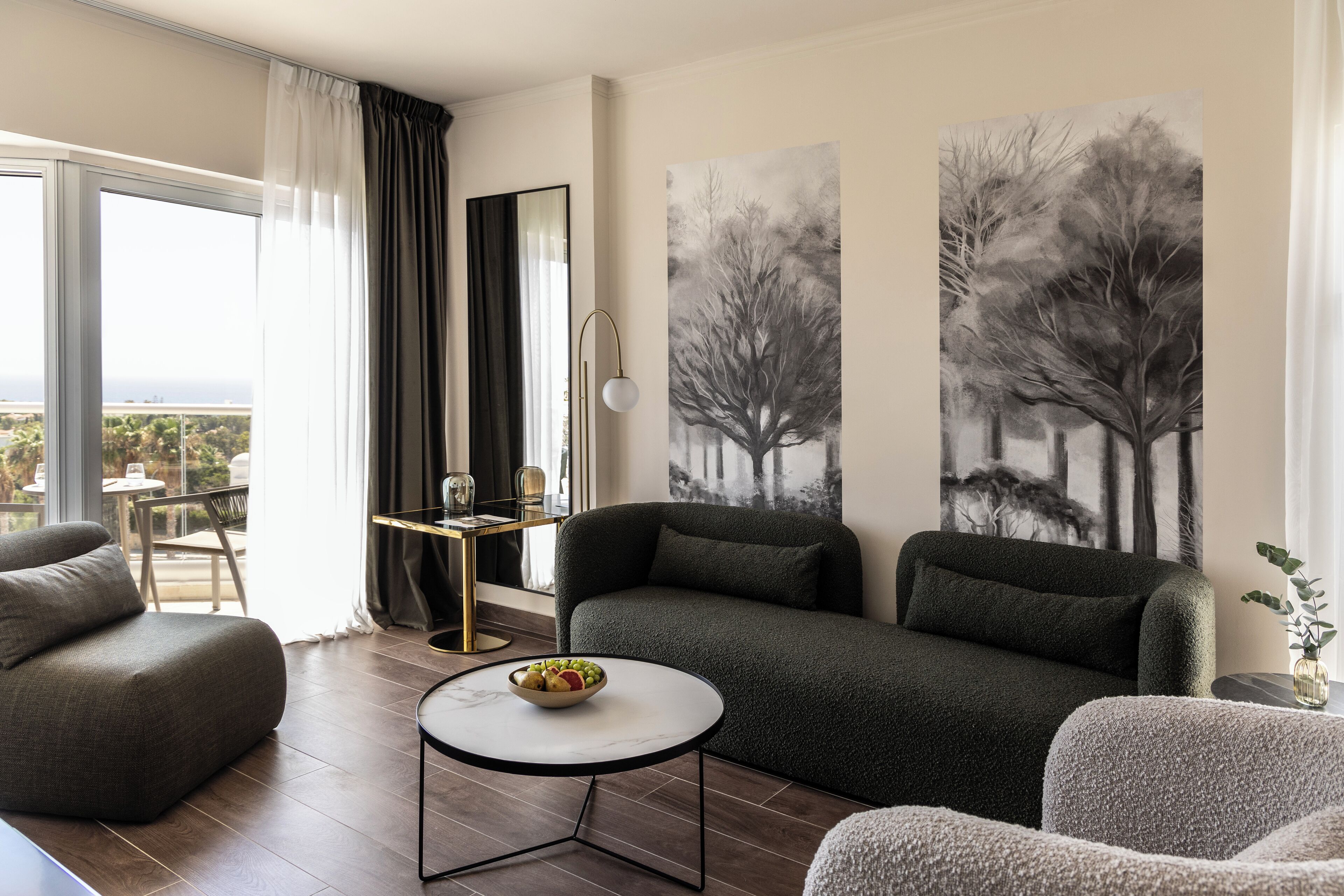 A modern living room with a sofa, armchair, and a small round table with a bowl of fruit. There is a large window with curtains, a painting of trees, and a view of the outdoors.