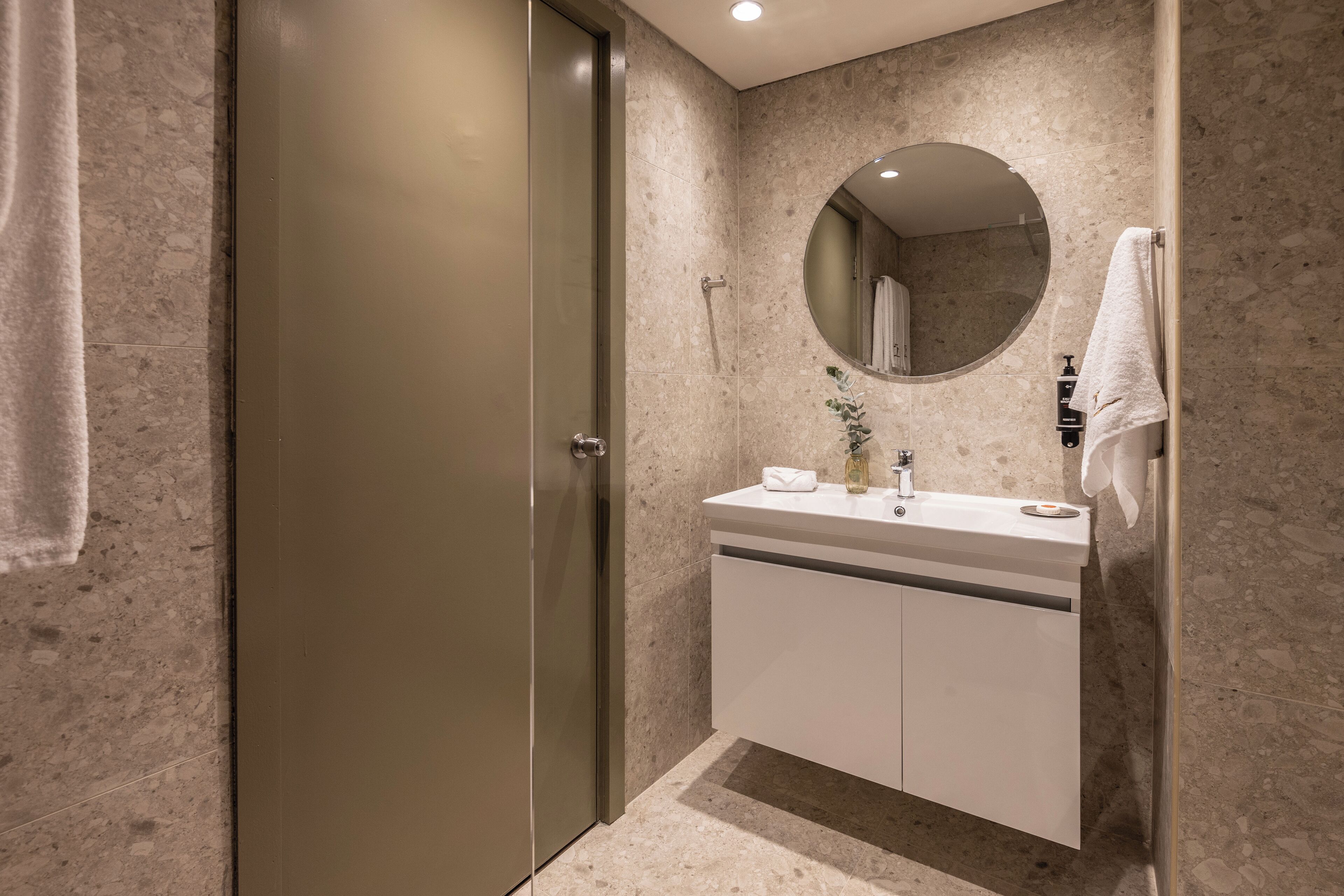 Un baño con pared de mármol, un espejo redondo, un lavabo con toalla y una puerta.