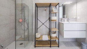 A modern bathroom with a glass shower enclosure, a wooden shelf unit with baskets, a white sink, and a mirror.