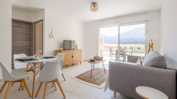 A modern living room with a sofa, dining table, and chairs, a television, and a view of the outdoors.