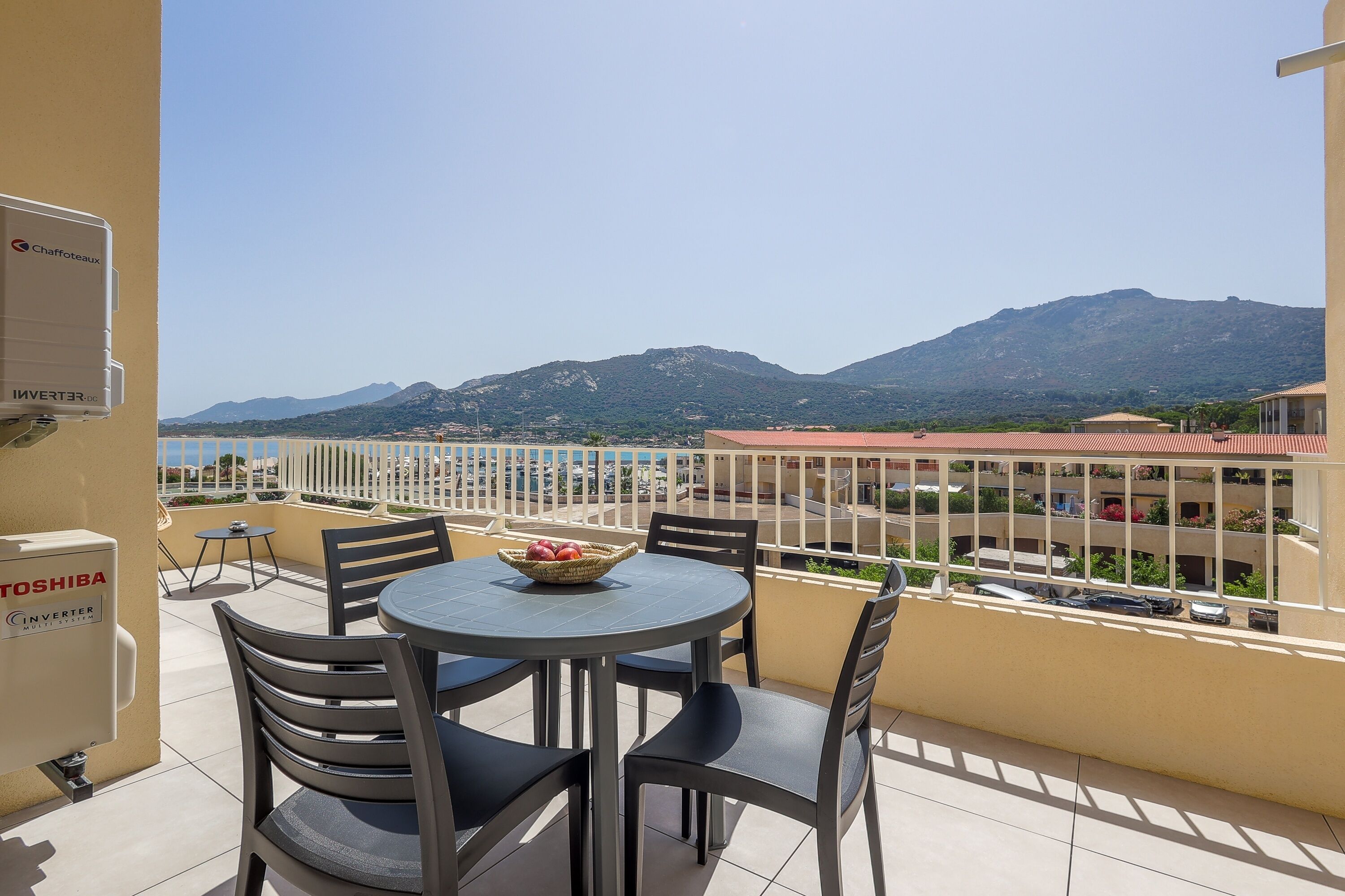 A balcony with a table and chairs, a basket of fruit, and a view of mountains and a beach.