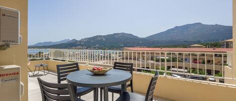 A balcony with a table and chairs, a basket of fruit, and a view of mountains and a beach.
