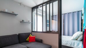A modern room with a grey sectional sofa, a bed with white bedding, and a large window with curtains. - Les Cormorans - Appt en rdc avec jardin (Benodet)