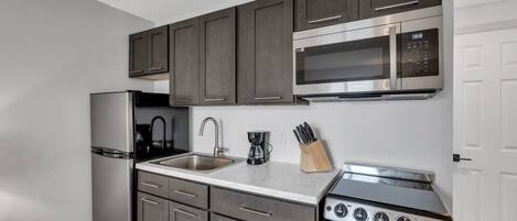 A compact kitchen with dark cabinets, a stainless steel refrigerator, a microwave, a sink, and a stove.