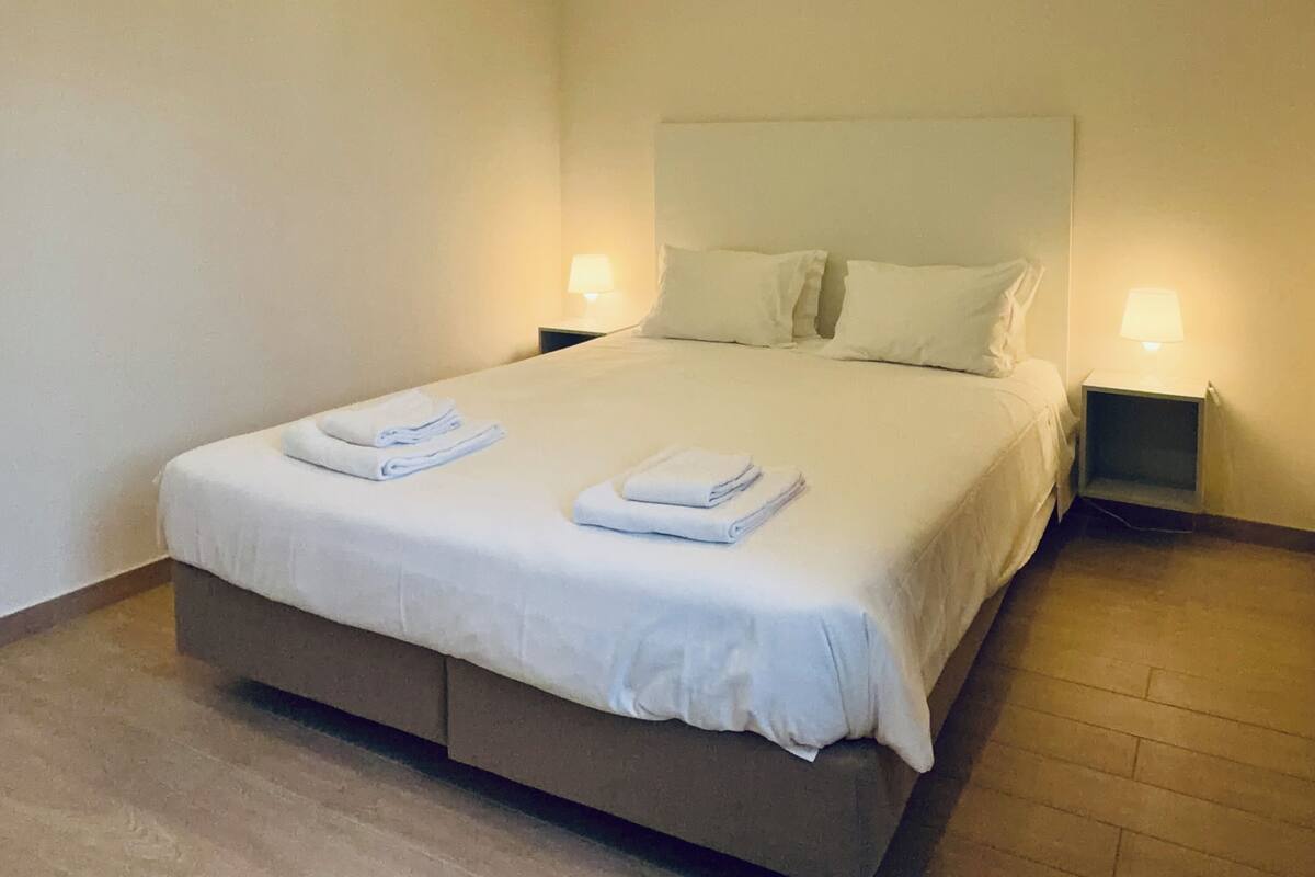 Un lit bien fait, avec du linge de lit et des serviettes blanches, deux lampes de chevet et un plancher en bois.