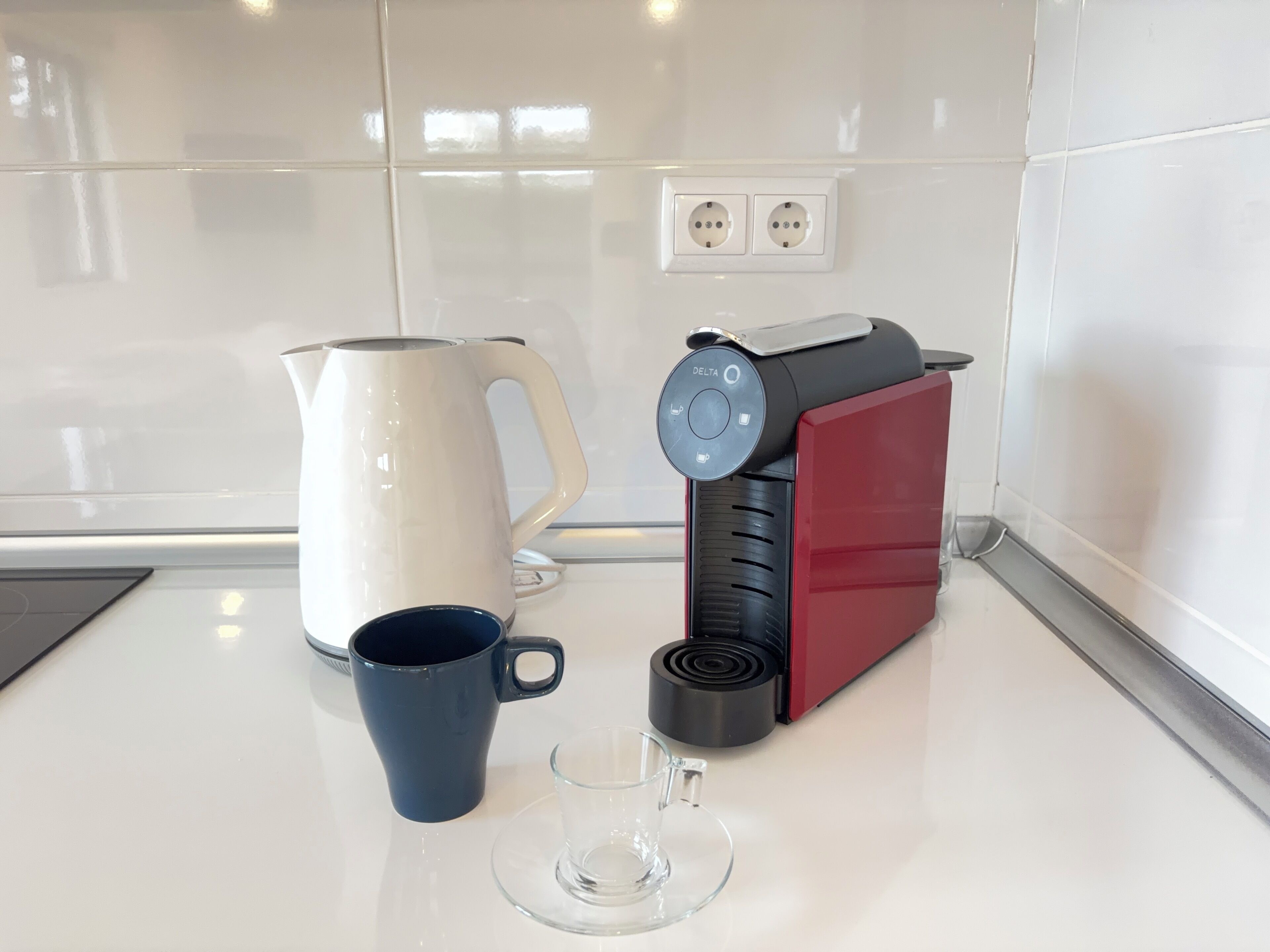 Une cuisine avec une machine à café, un pichet blanc, une tasse bleue et un verre transparent posés sur un plan de travail blanc.