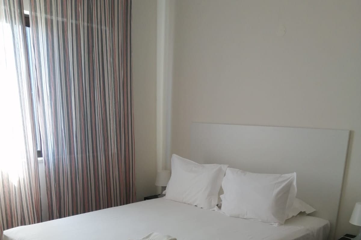 Un letto rifatto con lenzuola e cuscini bianchi, un asciugamano piegato sul letto, un pavimento in legno e una tenda a righe parzialmente tirata.