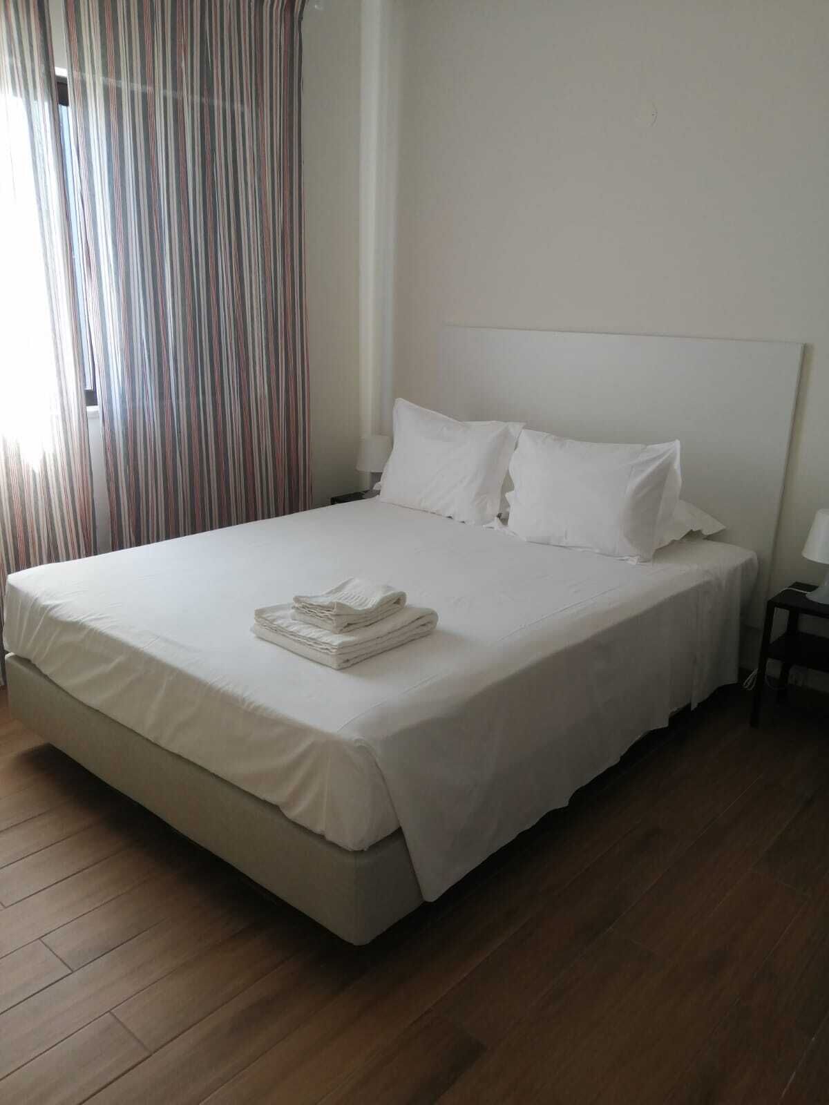 A neatly made bed with white linens and pillows, a folded towel on the bed, a wooden floor, and a striped curtain partially drawn.