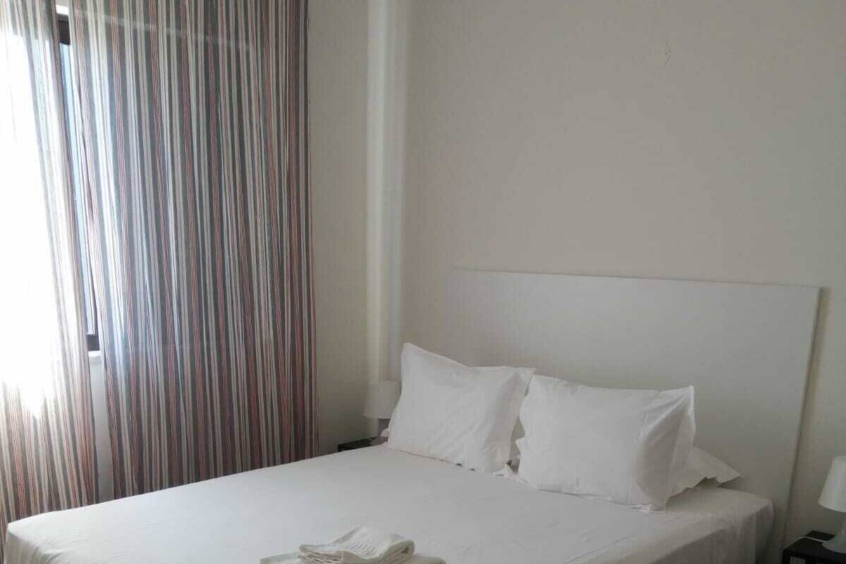 Un lit bien fait, avec du linge de lit blanc et des oreillers, une serviette pliée posée sur le lit, un plancher en bois et un rideau à rayures, partiellement tiré.